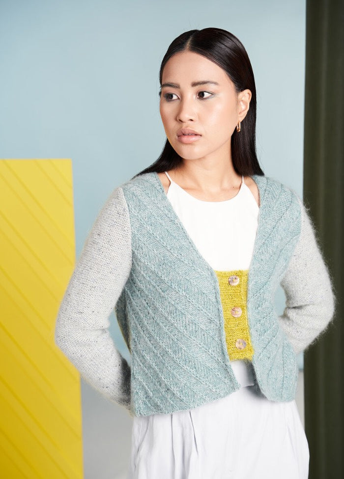 A woman with long dark hair looks to the side, wearing a textured blue cardigan over a white top with yellow accents. Background: light blue with a yellow panel.