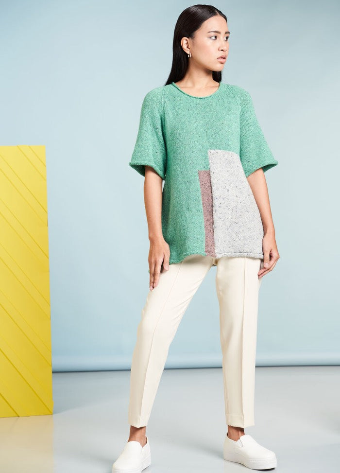 A woman in a green, color-blocked sweater and white pants stands against a light blue background. She wears white slip-on shoes, exuding a calm, modern vibe.