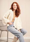 A woman with red hair sits on a chair, wearing a cozy white knit sweater with intricate patterns and a relaxed fit.