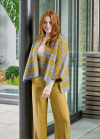 A woman with long red hair smiles, wearing a striped shawl and beige pants, standing indoors by glass windows. The scene feels modern and stylish.