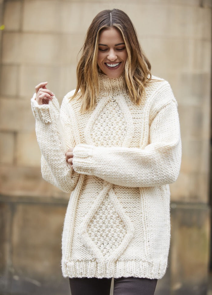 Smiling woman in a cozy, oversized ivory knit sweater with diamond patterns. Neutral background creates a warm, comfortable autumn vibe.