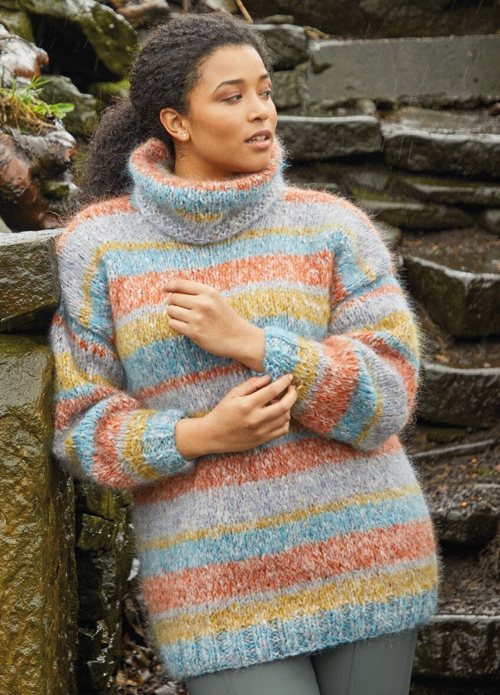 A woman in a cozy, striped turtleneck sweater stands by stone steps. The sweater features pastel blue, orange, and yellow hues, evoking warmth and comfort.
