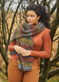 A woman stands among leafless trees, wearing a cozy, multicolored knit scarf and a rust orange sweater. The scene feels warm and serene.