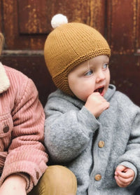 A toddler in a grey coat and mustard knit hat with a pom-pom, sits next to a person in a pink jacket, looking curious and playful against a wooden backdrop.