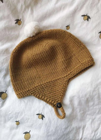 A knitted brown baby hat with ear flaps and a white pom-pom sits on a white blanket decorated with small yellow lemon patterns, creating a cozy feel.