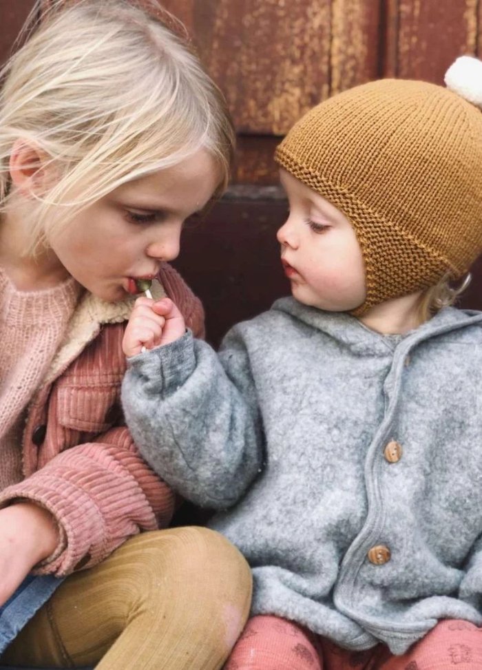 Two young children sit together. The older child wears a pink jacket and gently takes a treat from the younger child's hand, who is wearing a brown knit hat and grey coat. The mood is tender and caring.