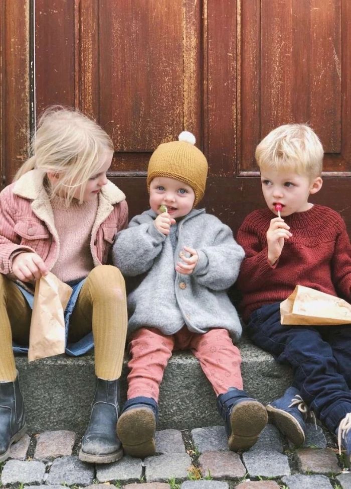 Three young children sit on a stone step against a wooden door, wearing cozy autumn clothes. They are enjoying lollipops and holding paper bags.