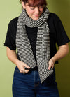 Person wearing a hand-knitted black and white striped scarf, styled with a black t-shirt and blue jeans, standing against a green background – cozy and casual knitwear look.