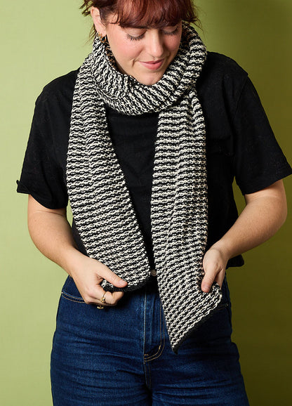 Person wearing a hand-knitted black and white striped scarf, styled with a black t-shirt and blue jeans, standing against a green background – cozy and casual knitwear look.