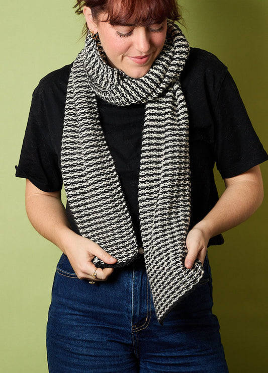 Person wearing a hand-knitted black and white striped scarf, styled with a black t-shirt and blue jeans, standing against a green background – cozy and casual knitwear look.
