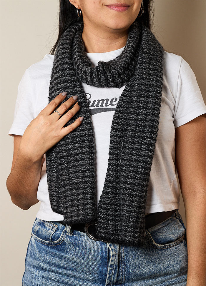 Person wearing a dark gray hand-knitted scarf paired with a white t-shirt featuring partial text and blue jeans – casual and cozy knitwear styling.