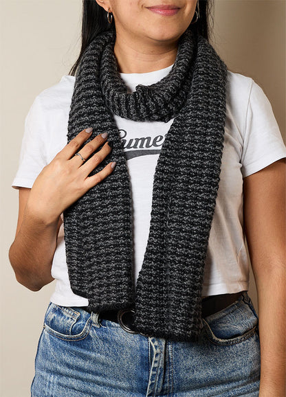 Person wearing a dark gray hand-knitted scarf paired with a white t-shirt featuring partial text and blue jeans – casual and cozy knitwear styling.