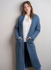 Woman in a cozy, blue knit cardigan over a white blouse, stands against a light gray wall. She has a relaxed pose with hands in pockets. Casual and stylish vibe.