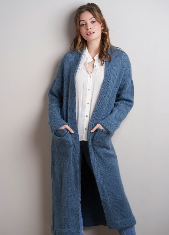 Woman in a cozy, blue knit cardigan over a white blouse, stands against a light gray wall. She has a relaxed pose with hands in pockets. Casual and stylish vibe.