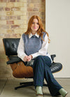 Young woman with long red hair sits on a modern black leather chair. She wears a light blue sweater vest, white shirt, and plaid pants, exuding a relaxed, stylish vibe.
