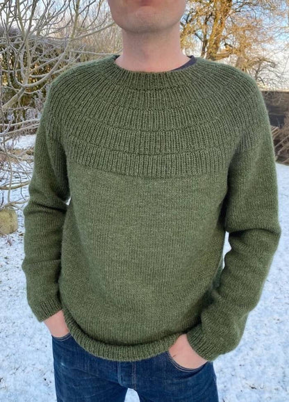 A person wearing a textured green sweater and blue jeans stands in a snowy outdoor setting. Bare trees and snow cover the ground, creating a serene winter atmosphere.