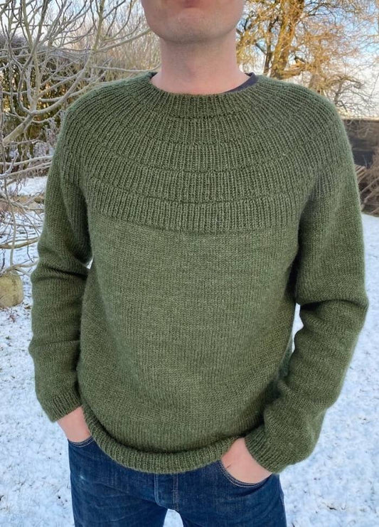 A person wearing a textured green sweater and blue jeans stands in a snowy outdoor setting. Bare trees and snow cover the ground, creating a serene winter atmosphere.