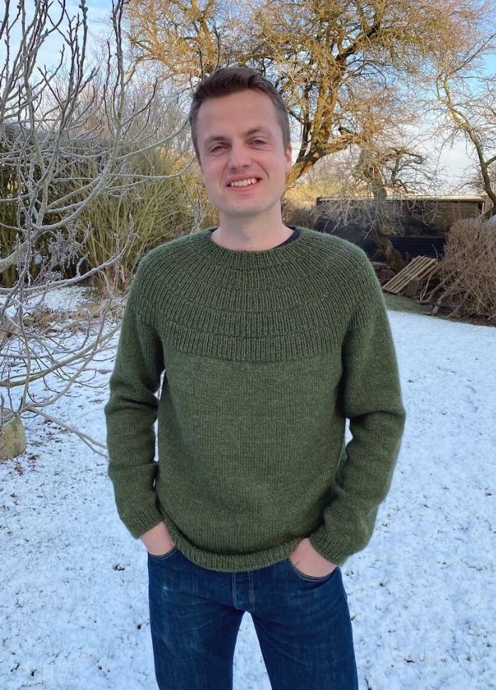 Man in a green sweater and jeans smiles in a snowy garden. Bare trees and a clear blue sky create a calm winter scene.