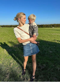 A woman in a white shirt and denim skirt holds a child in striped clothes in a sunny field. The mood is joyful and serene, with clear blue skies.