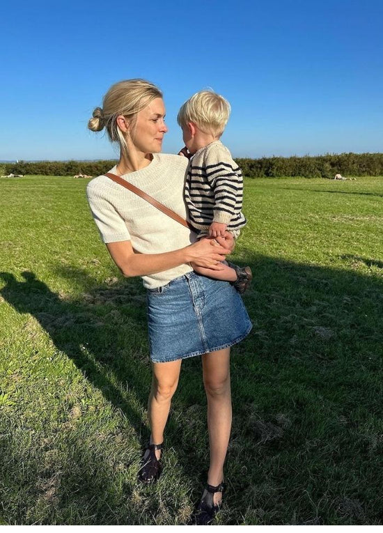 A woman in a white shirt and denim skirt holds a child in striped clothes in a sunny field. The mood is joyful and serene, with clear blue skies.