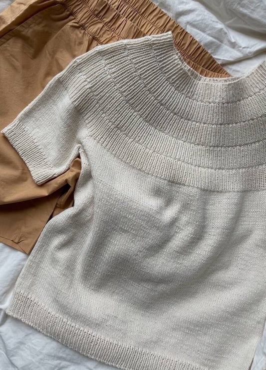 A cream knitted sweater with a textured yoke lies on a pair of tan, elastic-waist shorts. The clothing is displayed on a soft white background, evoking a cozy, casual vibe.