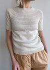 A person wearing a cream-coloured knit short-sleeve sweater with a textured, circular yoke pattern, paired with loose, light tan pants. The style appears casual and comfortable.