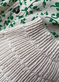 Close-up of a textured white knit fabric on a garment with a cream background and green leafy pattern. A dark button is visible, adding detail.