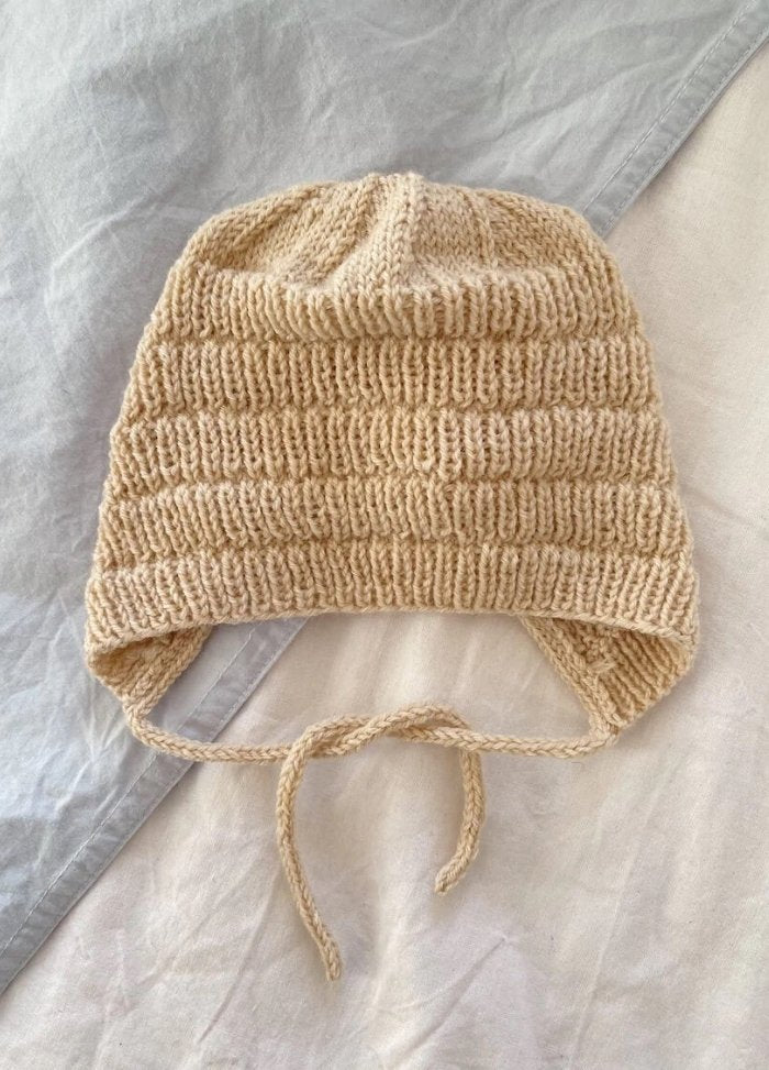 Knitted beige hat with earflaps and ties, placed on layered grey and cream fabric background. It evokes a cozy and crafted feel.