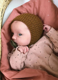 A baby in a mustard knit bonnet and pink outfit lies in a cozy setting. The baby has curious blue eyes and is wrapped in a soft pink blanket with tiny dots.