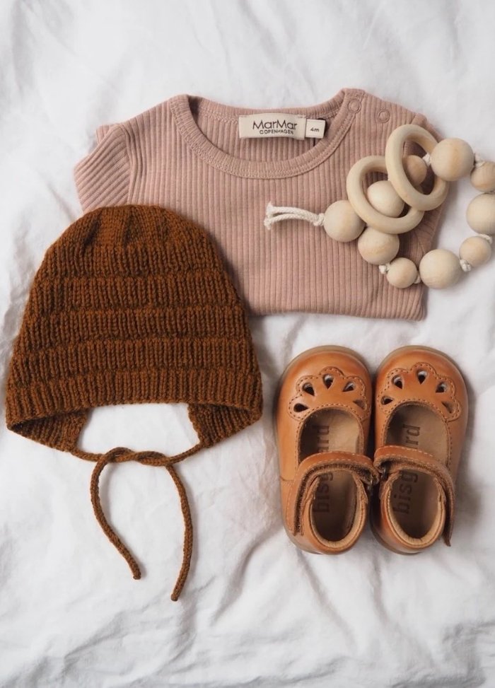 Baby essentials on a white background: a pink ribbed onesie, a brown knit bonnet, beige baby shoes with cut-out details, and a wooden teether.