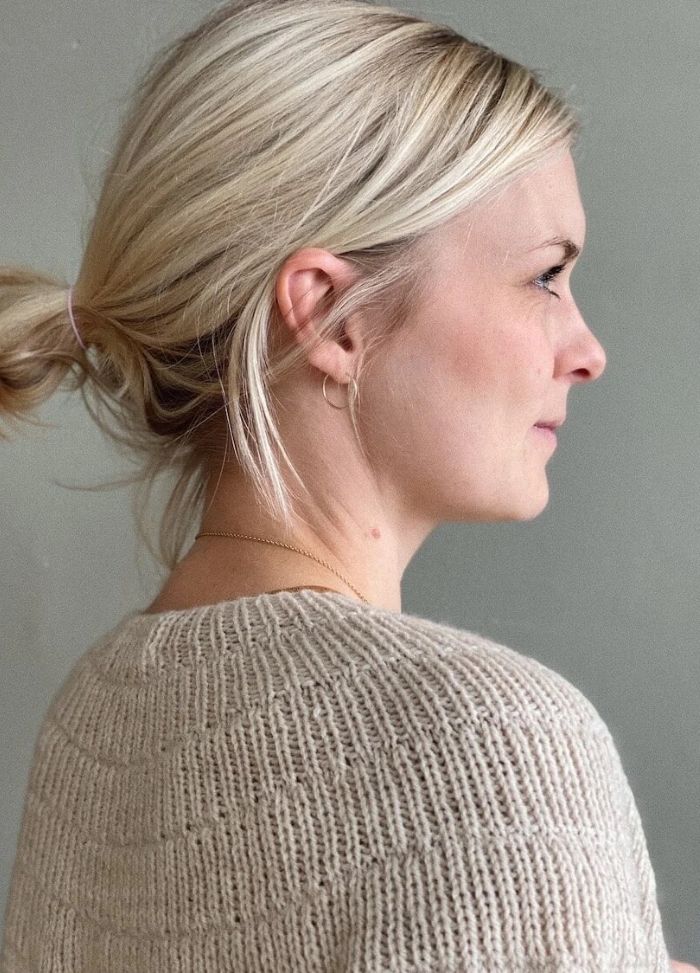 Profile of a woman with blonde hair tied in a bun, wearing a knitted beige sweater, looking calmly to the right against a gray background.