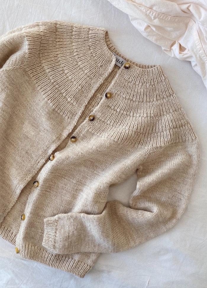 A cozy beige knit cardigan with a textured yoke and brown buttons is neatly laid on a soft white fabric background, exuding warmth and comfort.
