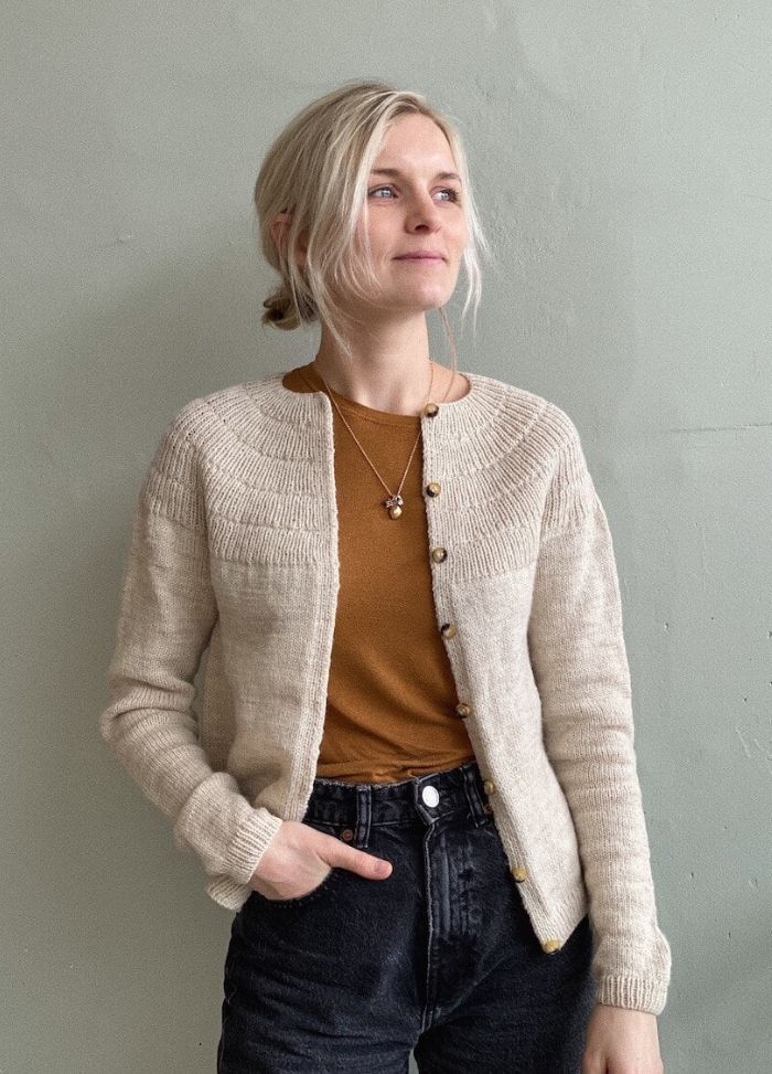 Woman with blonde hair stands against a light grey wall, wearing a cream cardigan, brown top, and black jeans. She looks to the side, appearing thoughtful.