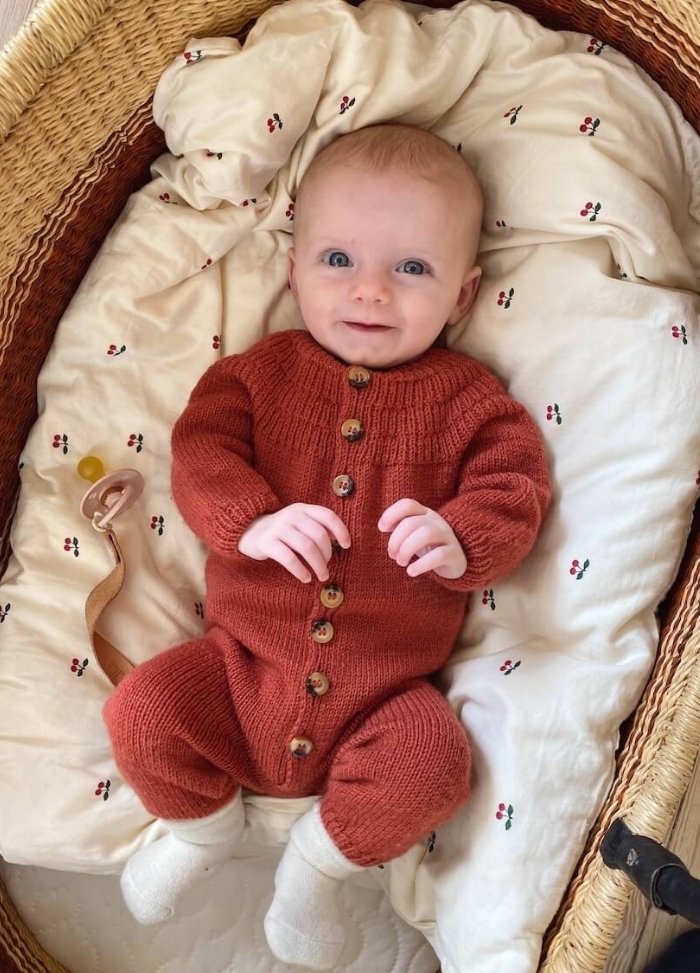 A smiling baby in a red knitted onesie lies on a patterned, cushioned blanket in a woven basket. The scene is cozy and cheerful, with a pacifier nearby.