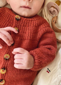 Close-up of a baby in a rust-coloured knitted onesie with decorative buttons, lying on a cream blanket with cherry patterns.