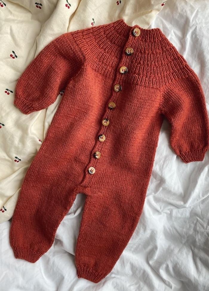 A cozy, rust-coloured knitted baby jumpsuit with wooden buttons lies on a crumpled, patterned white sheet featuring small cherries, evoking warmth and softness.