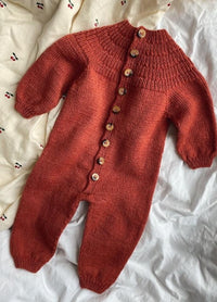 A cozy, rust-coloured knitted baby jumpsuit with wooden buttons lies on a crumpled, patterned white sheet featuring small cherries, evoking warmth and softness.