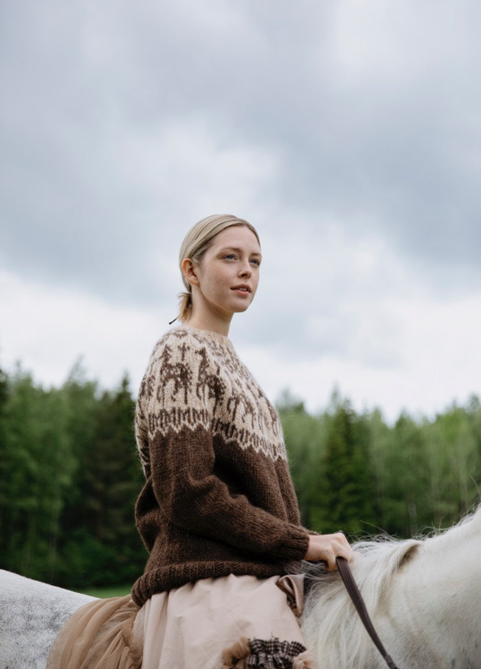 Anna Huolia Hevosen Sweater: brown knit with horse motifs around the yoke, worn while riding a white horse in a forested setting.