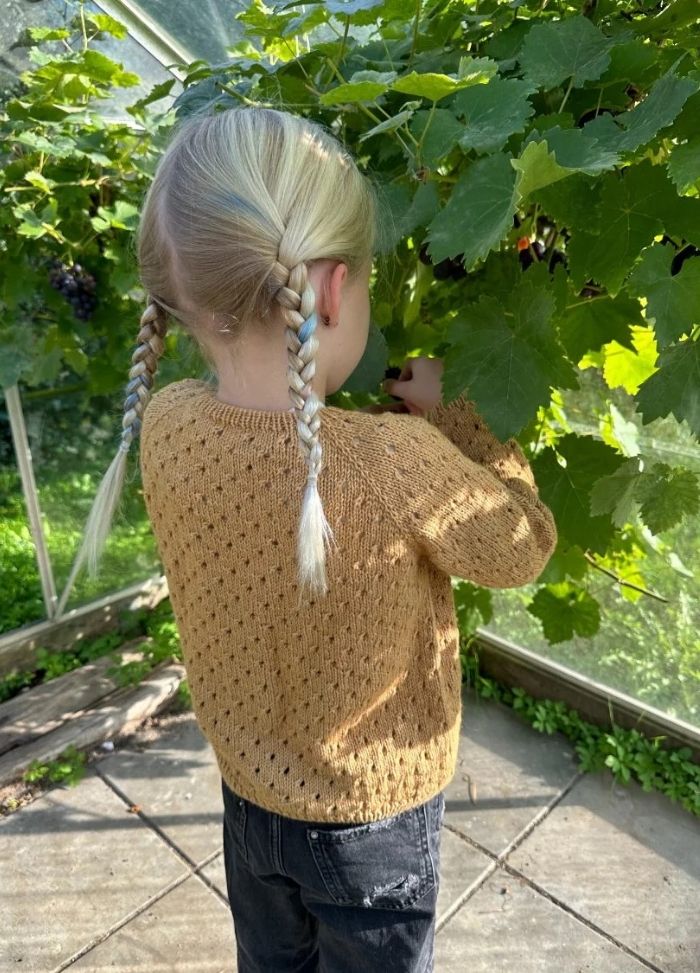 A child with braided hair, wearing a yellow cardigan and black jeans, stands in a sunlit greenhouse picking grapes. The scene is calm and natural.