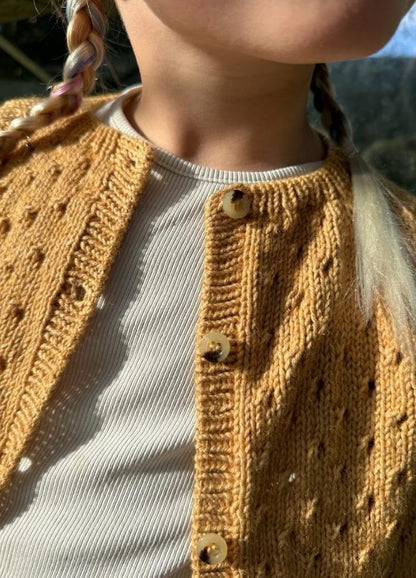 Close-up of a person wearing a textured mustard yellow cardigan over a cream ribbed shirt. Braided hair strands are visible, with sunlight casting soft shadows.