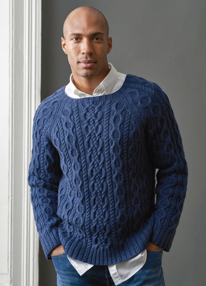 A man dressed in a blue sweater and jeans poses confidently, showcasing a relaxed style.
