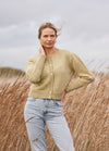 Woman stands in a field of tall grass, wearing a beige knit cardigan and jeans. Her hair is gently blown by the wind, under a cloudy sky. Calm and serene mood.