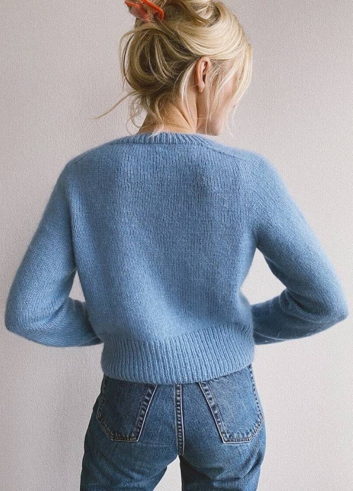 A person with blonde hair pinned up with a red clip is wearing a soft blue sweater and denim jeans, standing with their back to the camera.