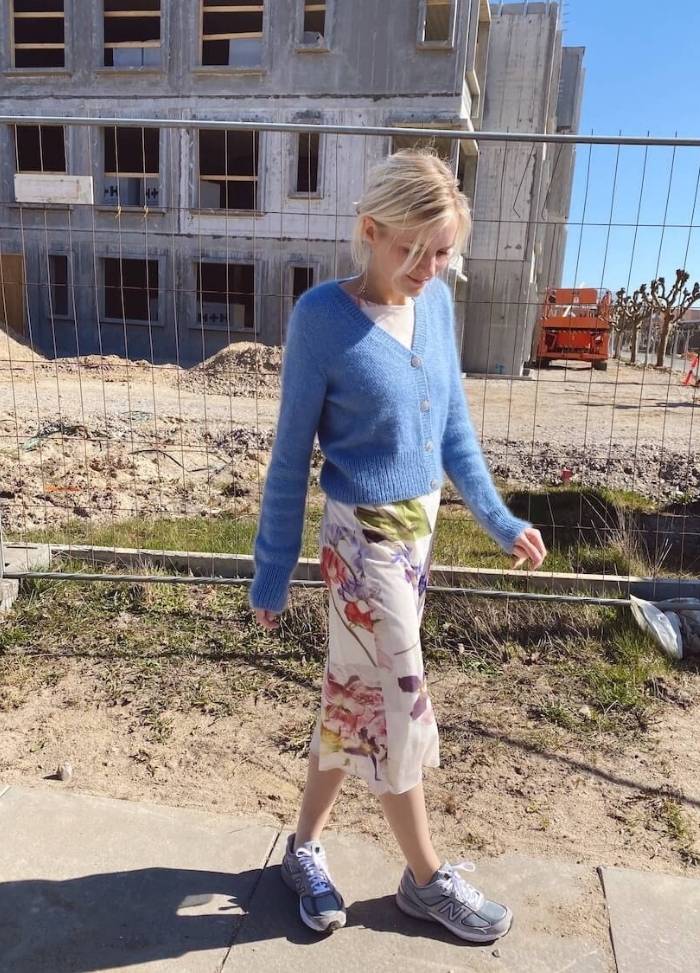 Young woman walks on a sunny day wearing a blue cardigan and floral skirt, near a construction site with a partially built concrete building in the background.