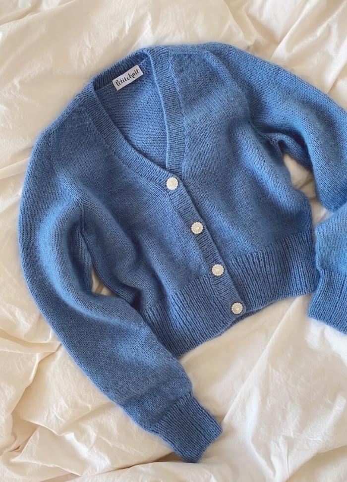 Blue knitted cardigan with a deep V-neck, white buttons, and long sleeves, lies on a cream-coloured fabric, conveying softness and warmth.