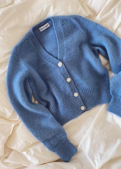 Blue knitted cardigan with a deep V-neck, white buttons, and long sleeves, lies on a cream-coloured fabric, conveying softness and warmth.