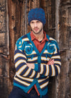 Man in a colorful striped cardigan and blue knit hat leans against a rustic wooden wall. His expression is contemplative, adding an introspective mood.