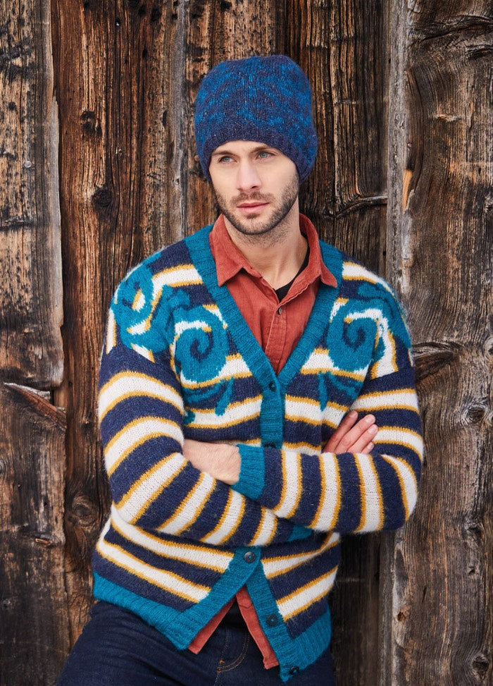 Man in a colorful striped cardigan and blue knit hat leans against a rustic wooden wall. His expression is contemplative, adding an introspective mood.