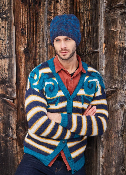 Man in a colorful striped cardigan and blue knit hat leans against a rustic wooden wall. His expression is contemplative, adding an introspective mood.