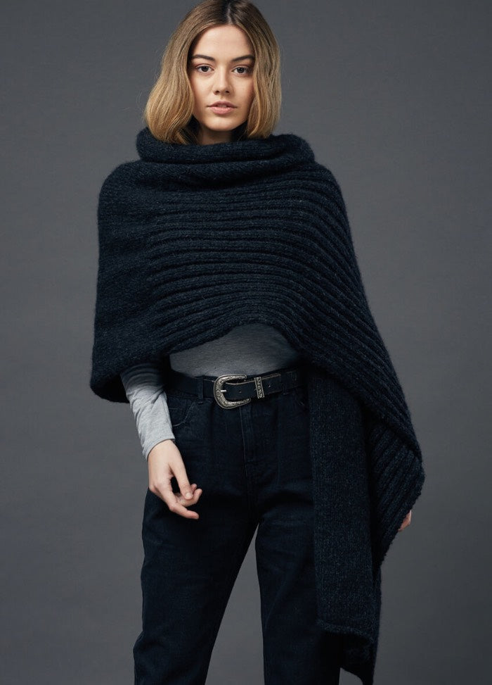 A woman stands against a gray background, wearing a dark knit shawl, gray top, and black pants. Her expression is calm, conveying a relaxed mood.
